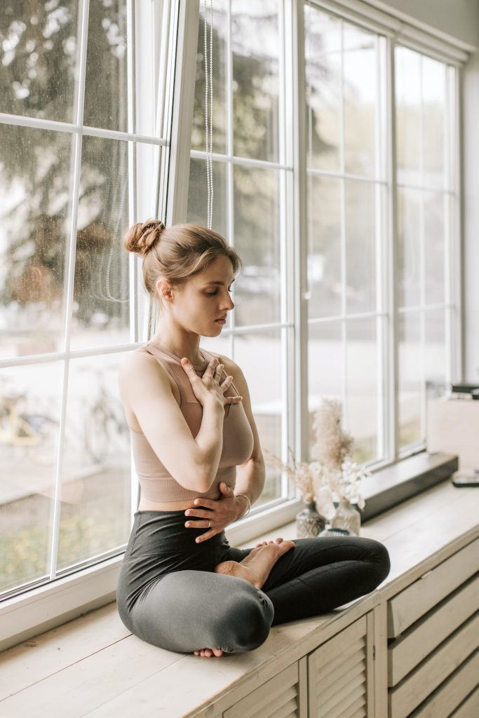 Woman in Beige Tank Top Sitting Near the Glass Windows while Meditating